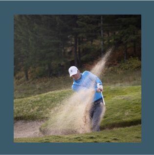 A person in a blue jacket and white cap is skillfully hitting a golf ball out of a sand trap on a scenic golf course, surrounded by the serene beauty of Suncadia.