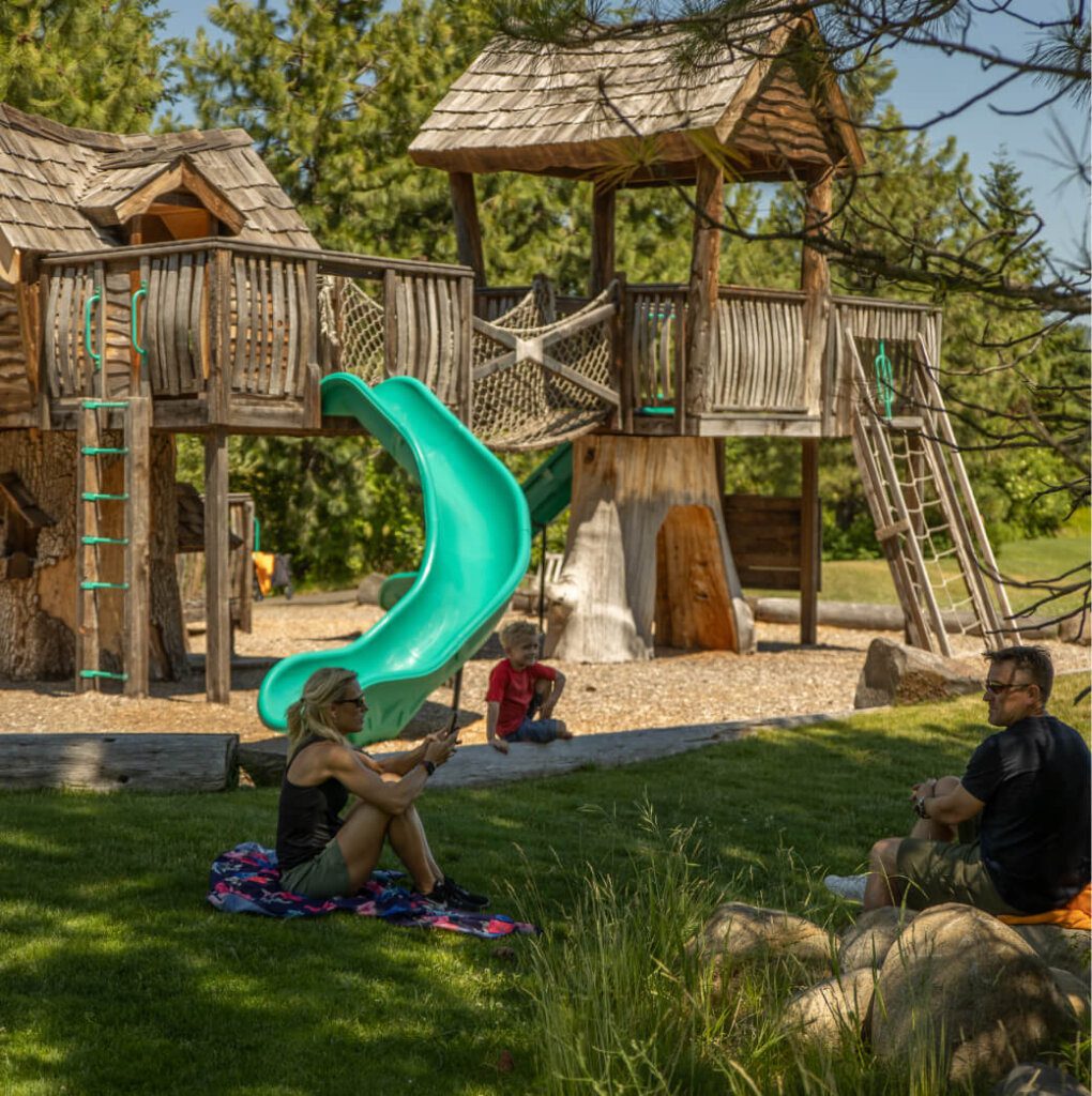 Adults and a child relax near a wooden playground structure with a green slide in a grassy park, enjoying the serene atmosphere reminiscent of Suncadia Resort in Washington. It's the perfect spot to unwind before exploring Cle Elum restaurants or checking out Suncadia real estate opportunities nearby.