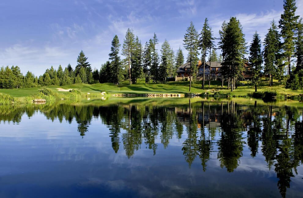 A serene golf course with a tranquil pond reflects tall evergreens, offering peace near the Suncadia real estate. The distant clubhouse rests under a clear blue sky, while nearby Cle Elum restaurants await your post-game indulgence.