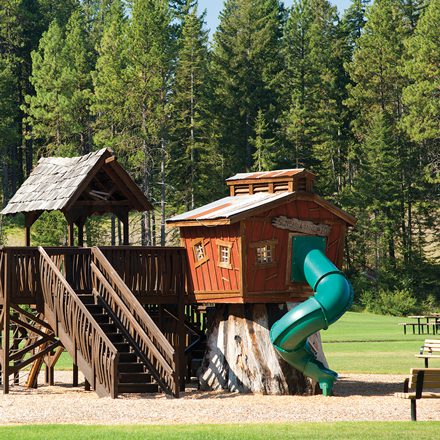 A wooden playground structure with a green spiral slide stands proudly in an open grassy area at Suncadia Resort, surrounded by lush trees that add to the serene beauty of Washington's landscape.