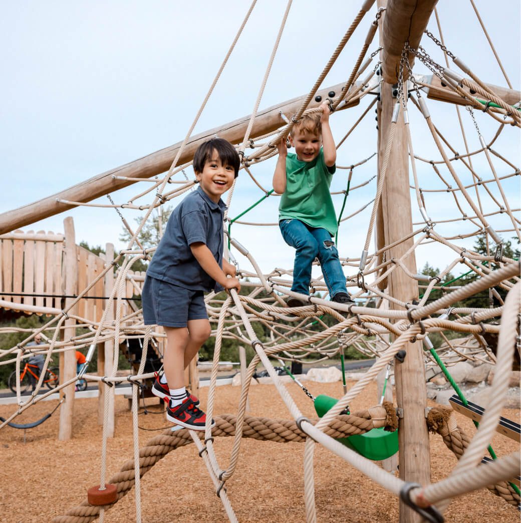 Two children are climbing on a rope playground structure with wooden poles at the scenic Suncadia Resort. The background showcases more playground equipment and lush trees, offering a perfect blend of adventure and natural beauty that epitomizes the charm of Suncadia.