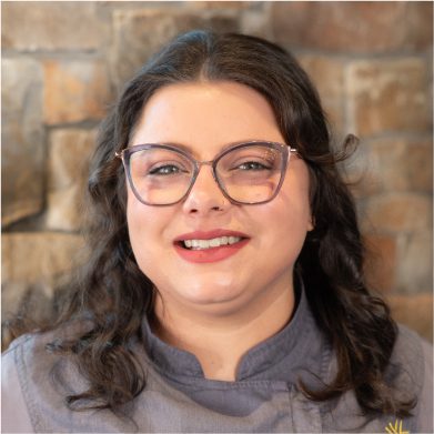 A person with long dark hair and glasses smiles at the camera, standing in front of a stone wall at Suncadia Resort in Washington.