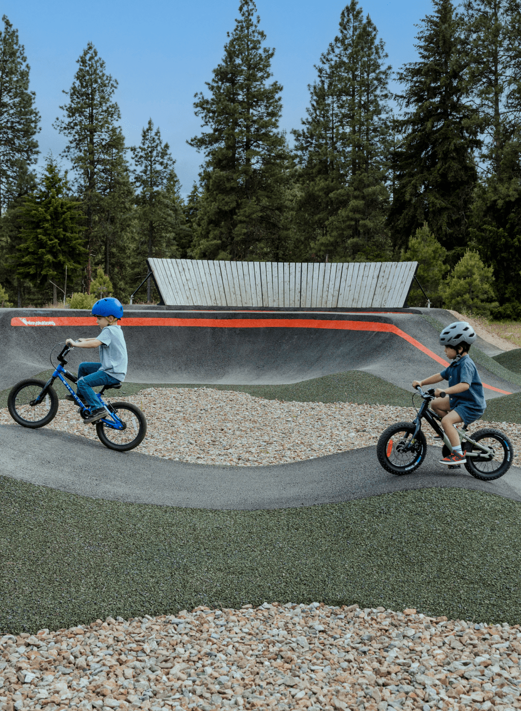 Two children wearing helmets ride bicycles on a paved pump track in a wooded area, with trees and a wooden fence in the background.