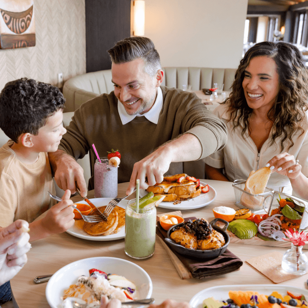 A family of four enjoying a meal at a restaurant, with various dishes and drinks on the table. The scene shows a cozy dining atmosphere with smiles and conversation.