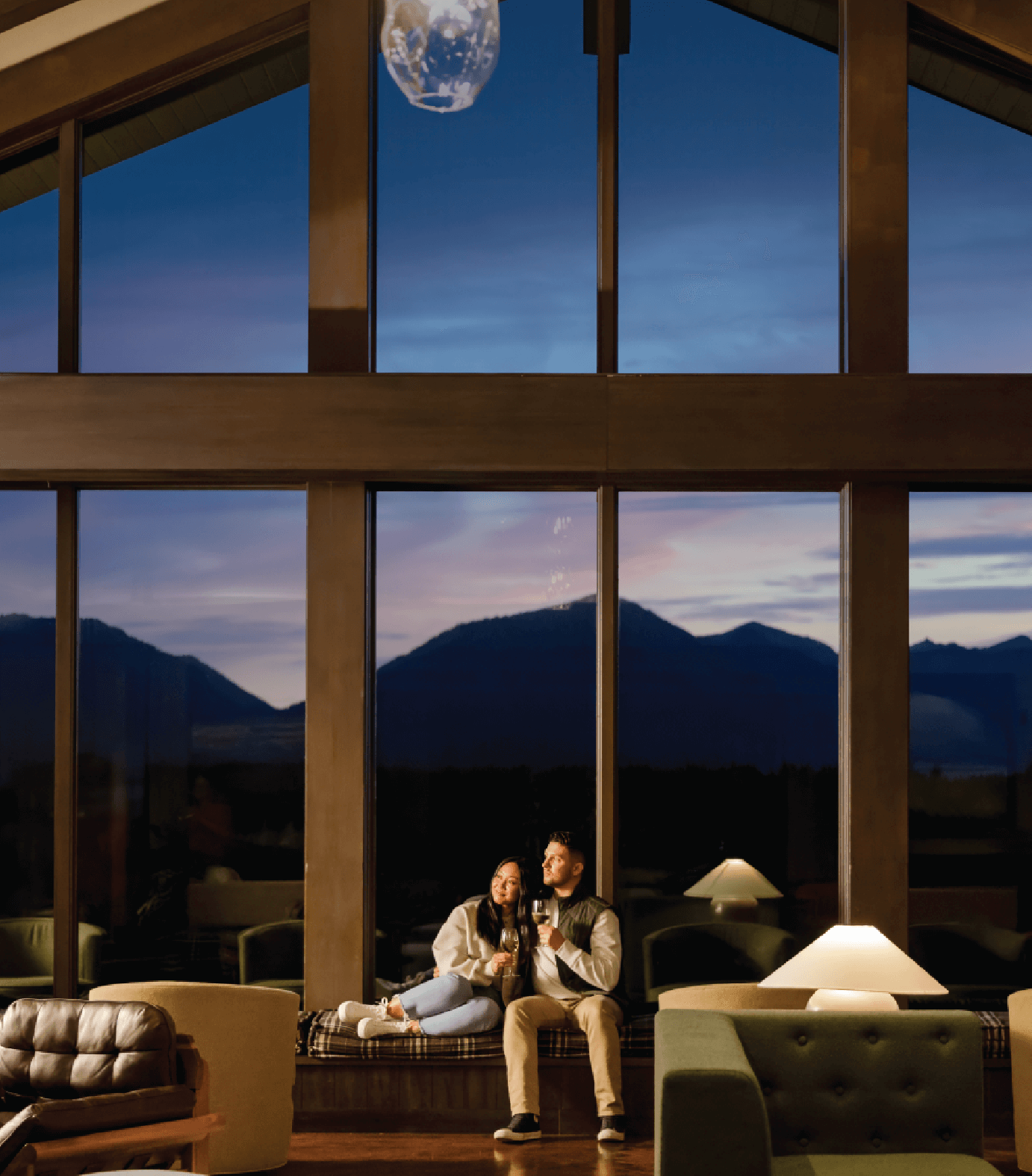 A couple sits in a cozy living room with large windows, watching the sunset over distant mountains.