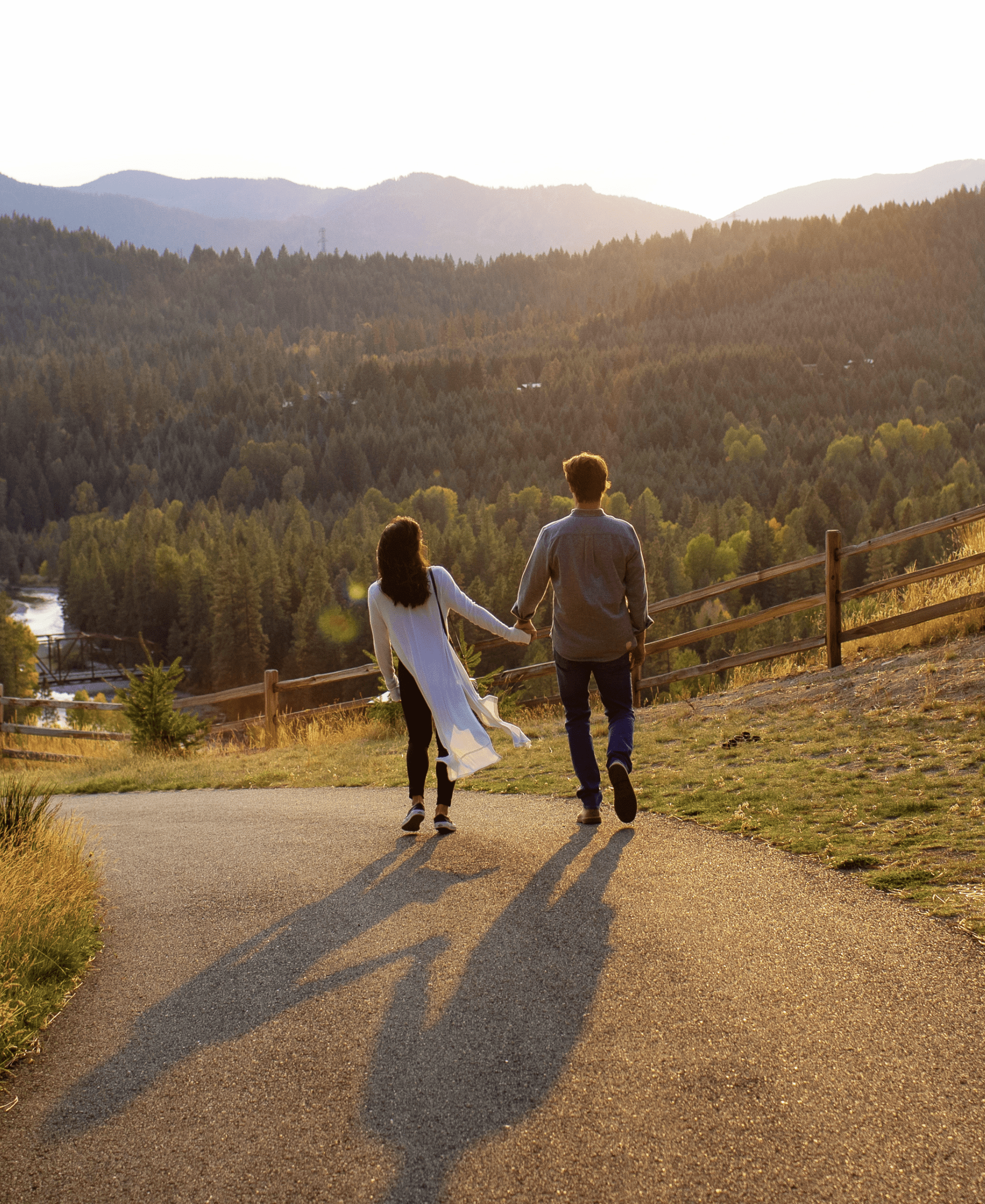 A couple walks hand in hand down a path with a scenic view of mountains, trees, and a setting sun.