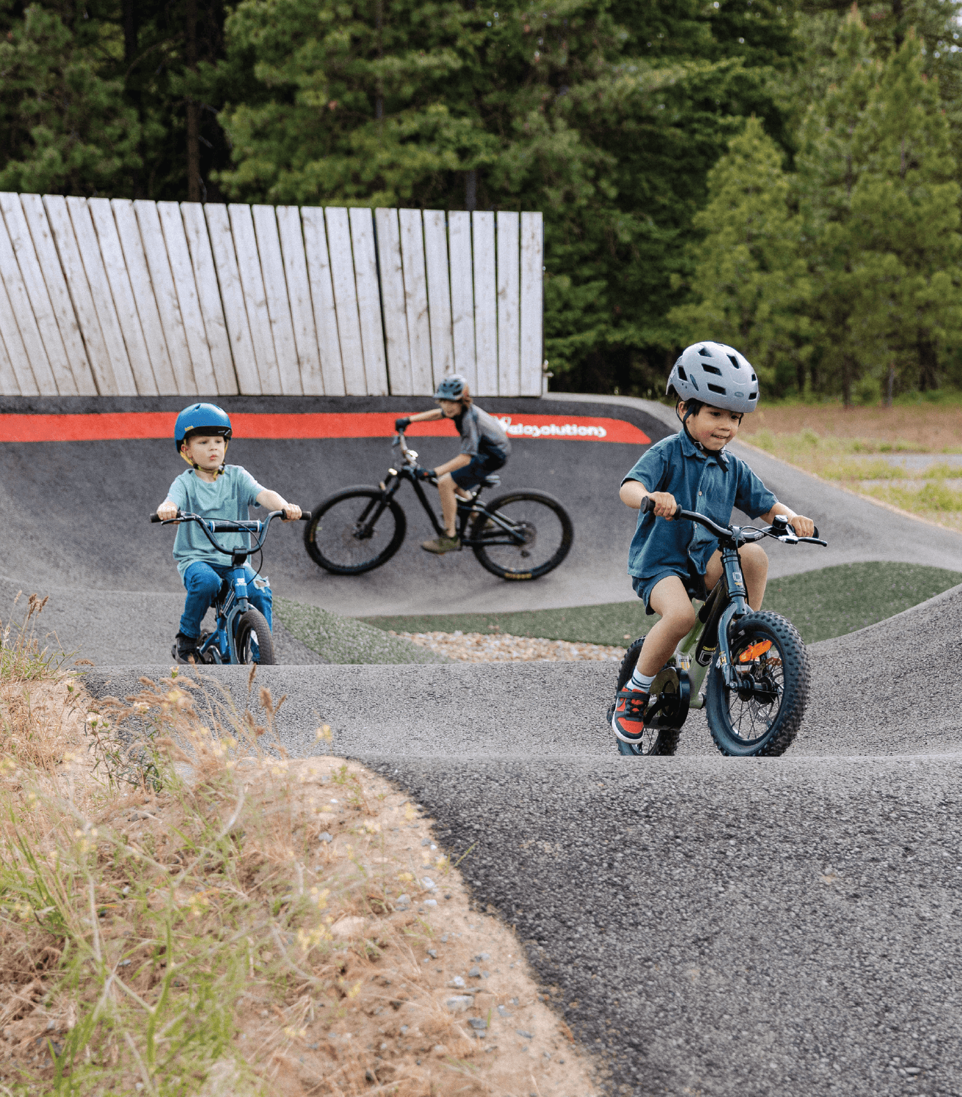 Children wearing helmets ride bikes on a pump track, followed by an adult in the background.
