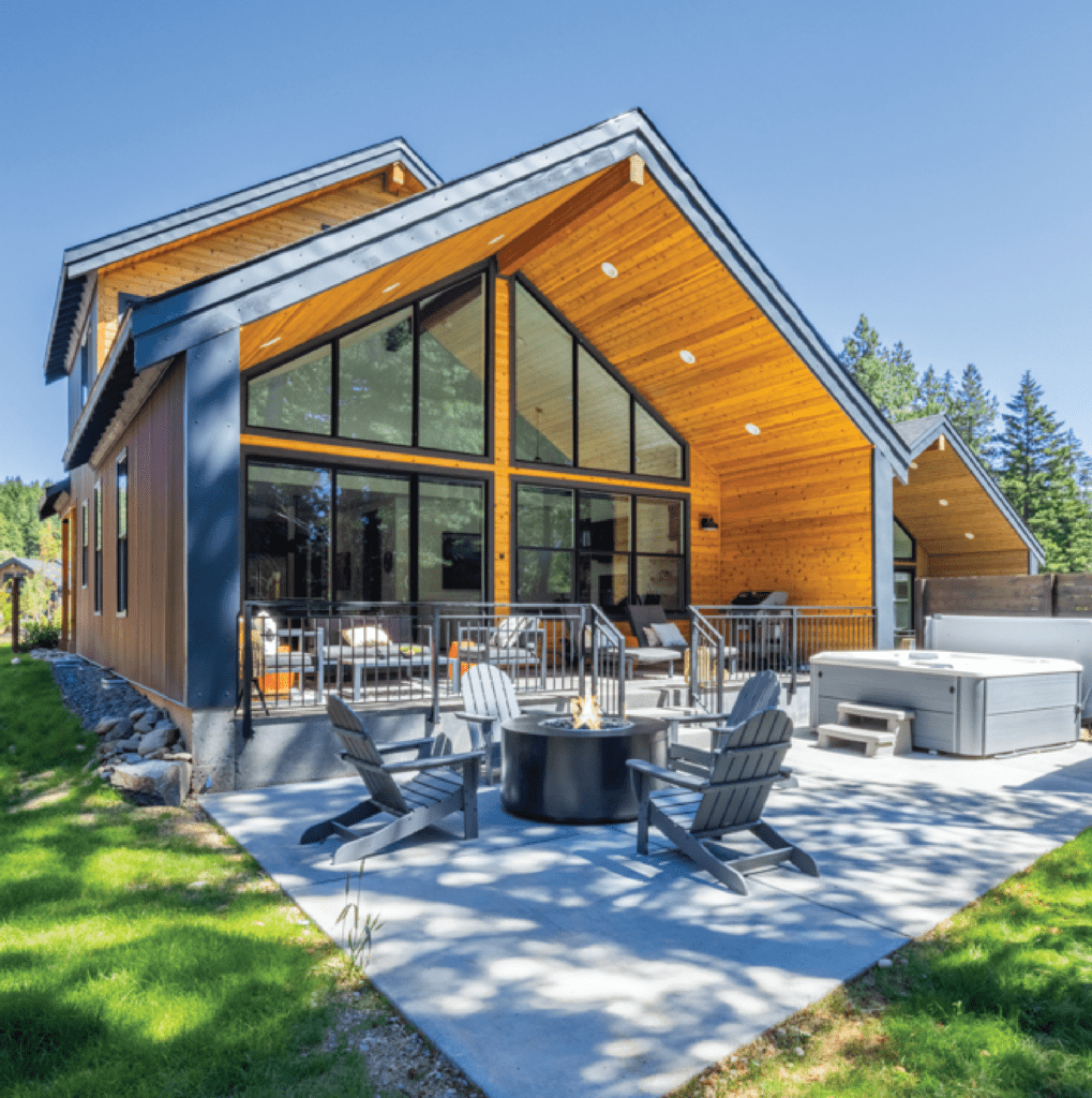 Modern wooden house with large windows, a patio with Adirondack chairs around a fire pit, and a hot tub. Lush green lawn and trees in the background.