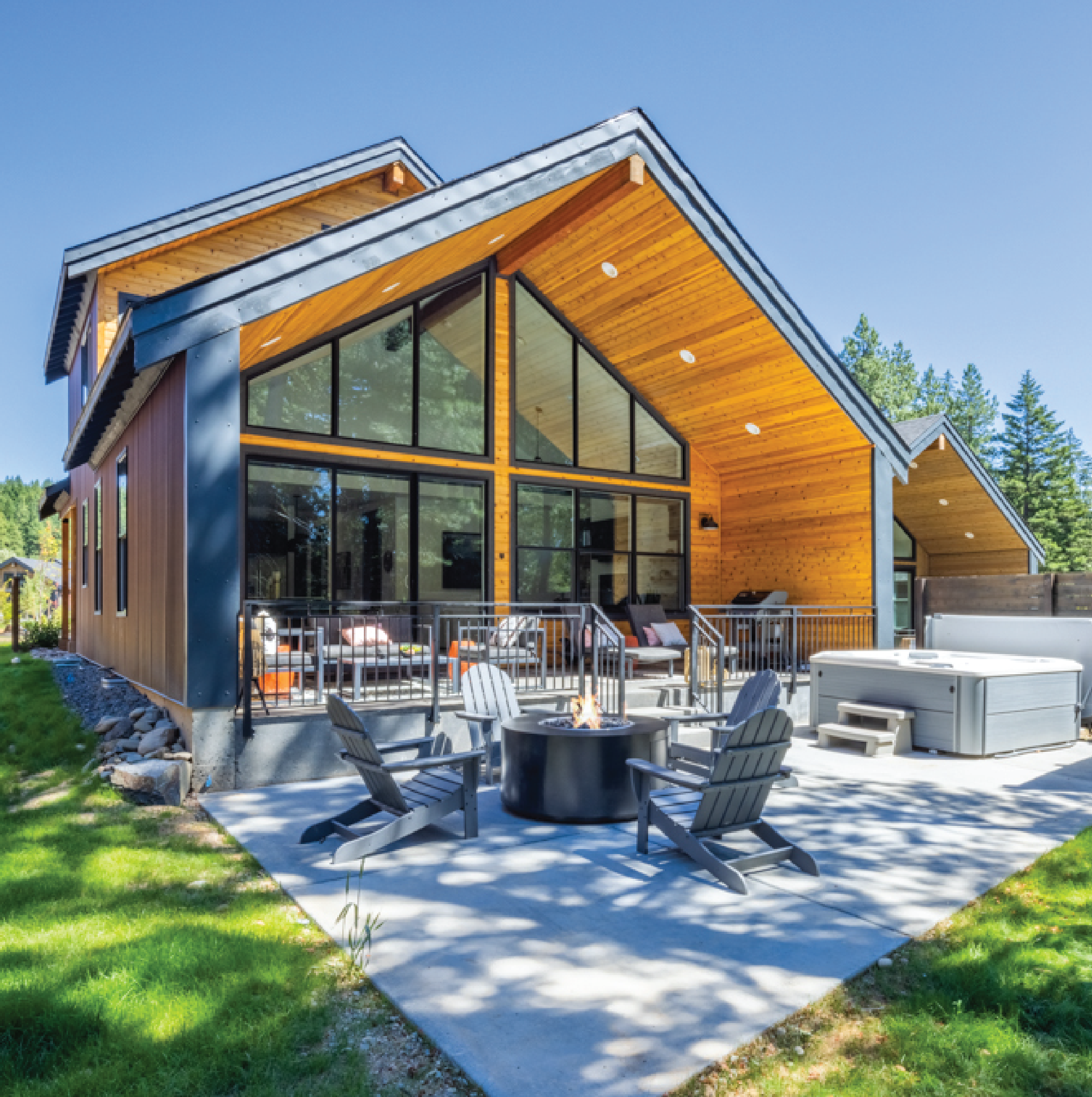 Modern wooden house with large windows, a patio with Adirondack chairs around a fire pit, and a hot tub. Lush green lawn and trees in the background.