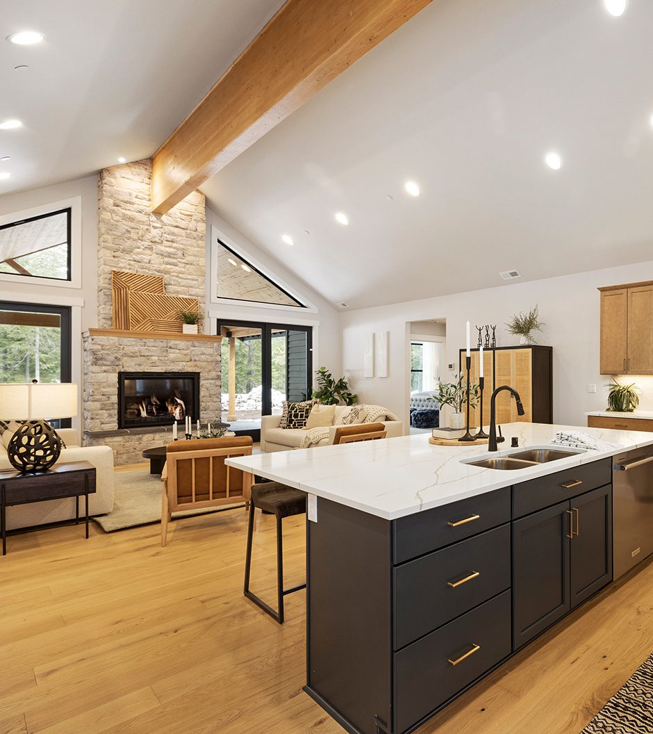 Open-concept living area featuring a modern kitchen with an island, a stone fireplace, wood flooring, and large windows letting in natural light.