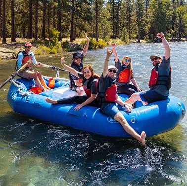 Six people wearing life jackets sit and pose on a blue inflatable raft on a river, surrounded by trees on a sunny day.
