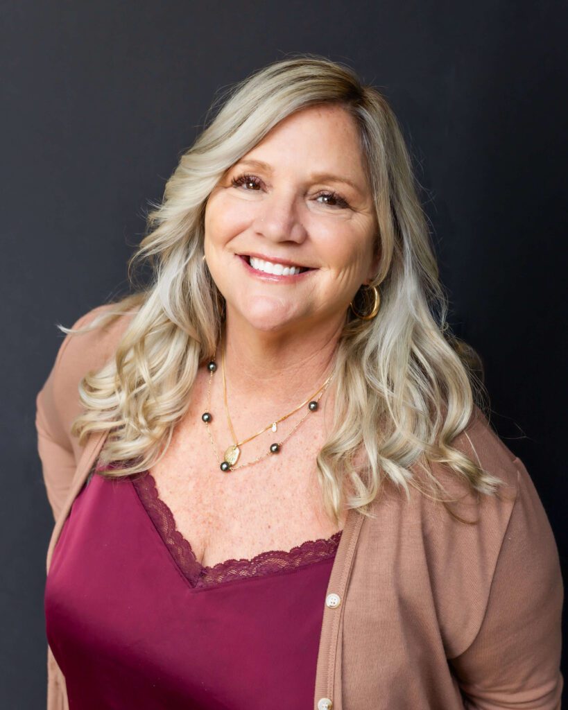 A woman with long blonde hair smiles at the camera, wearing a maroon top, tan cardigan, and layered necklaces against a plain dark background.