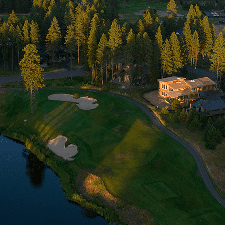 Aerial view of a golf course with sand traps, a water hazard, surrounding trees, and houses near the green in the evening sunlight.