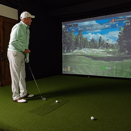 A man stands on artificial turf holding a golf club, preparing to hit a ball toward a virtual golf course displayed on a large screen.