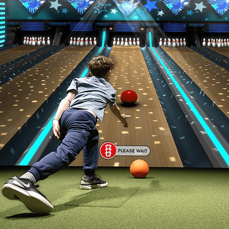 A child bowls a red ball toward pins in a neon-lit bowling alley, with another orange ball nearby and a “PLEASE WAIT” sign on the lane.