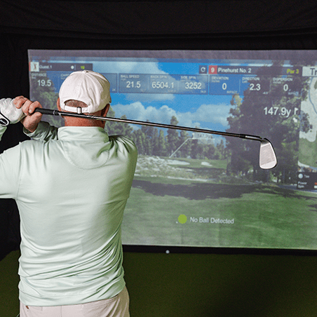 A person in a light shirt and cap practices golf indoors, facing a virtual golf simulator screen displaying course data and the message “No Ball Detected.”.