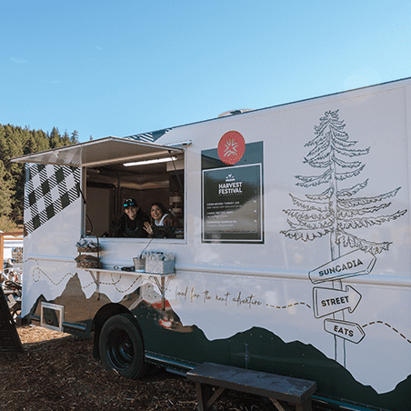 A food truck with "Suncadia Street Eats" branding is parked outdoors; two people are visible inside, serving at the window, with trees and a blue sky in the background.