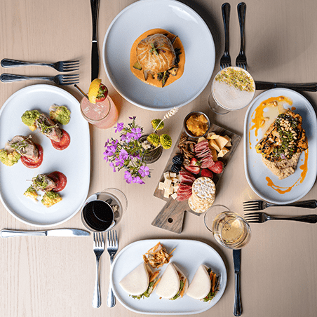 Overhead view of a table set with various plated dishes, charcuterie board, drinks, and cutlery, with a small floral centerpiece.