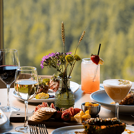 A table set with wine, cocktails, assorted appetizers, crackers, cheese, fruit, and a vase of wildflowers, with a forest view in the background.