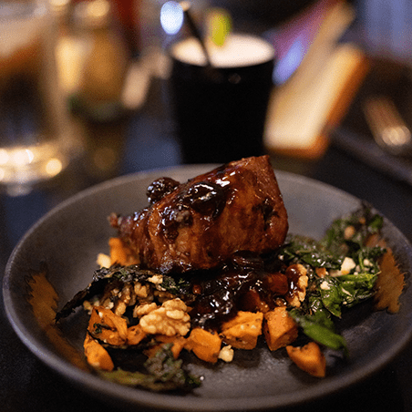 A plate with a piece of glazed meat on top of chopped sweet potatoes, wilted greens, and walnut pieces, served on a dark table.