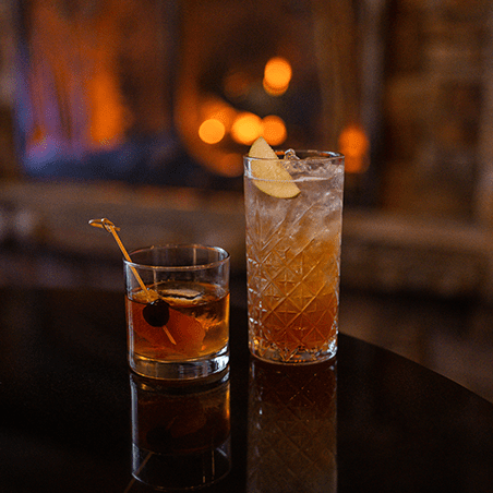 Two cocktails on a dark table: one in a short glass with an orange twist and cherry, the other in a tall glass with ice and a lemon slice. Warm lights are blurred in the background.