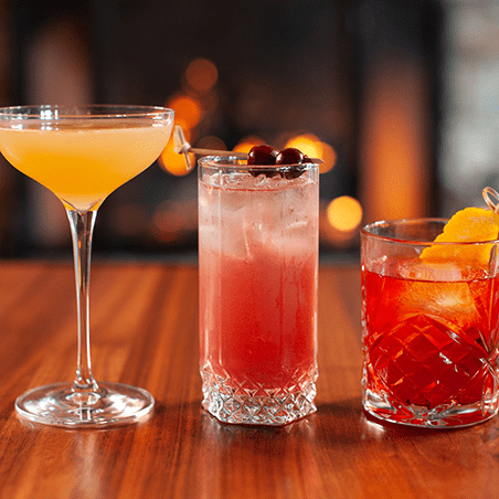 Three cocktails on a wooden table: a yellow drink in a coupe glass, a pink drink with cherries in a highball glass, and an orange drink with an orange peel in a rocks glass.