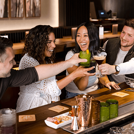 Four people sitting at a bar clink glasses and smile, with drinks and barware visible in front of them.