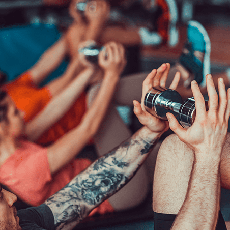 People lying on exercise mats holding dumbbells above their chests, with focus on a tattooed arm in the foreground.