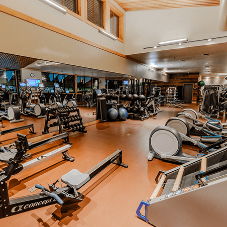Spacious gym with exercise machines, free weights, rowing machines, stability balls, and large mirrors; well-lit with high ceilings and wooden accents.