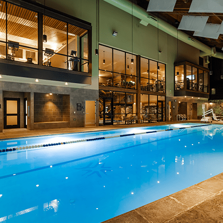 Indoor swimming pool with clear water, marked lanes, and modern gym facilities visible through large windows on the upper and lower levels.