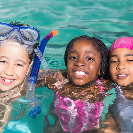 Three children in swimwear smile in a pool; one wears goggles and a snorkel, another has braided hair, and the third wears a pink swim cap.