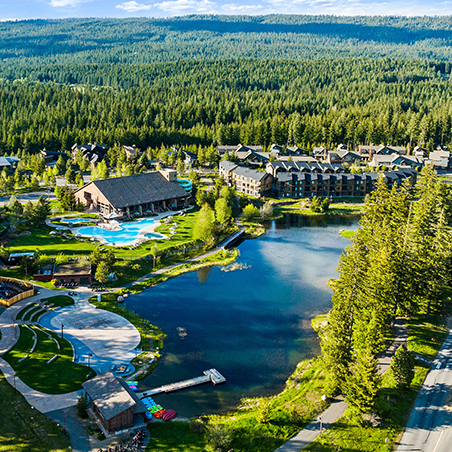 Aerial view of a resort complex with outdoor pools, a lake, surrounding trees, and forested hills in the background under a partly cloudy sky.