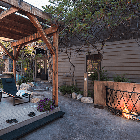 Outdoor patio area with wooden pergola, lounge chair, fire pit, and landscaped garden beside a modern rustic building.