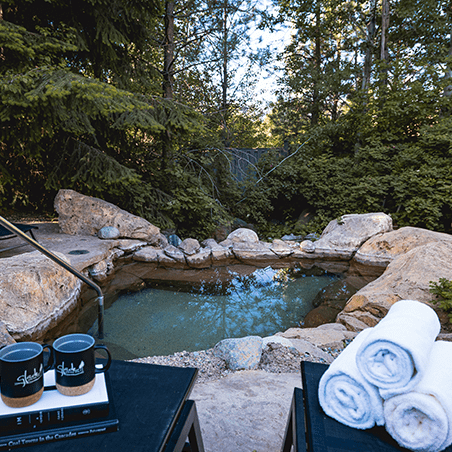 Outdoor hot spring pool surrounded by rocks and trees, with two towels on a chair and two mugs on a table in the foreground.