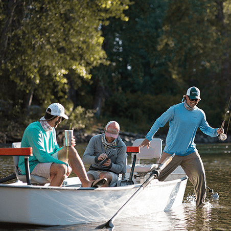 Three people are fishing on a small boat in a calm, wooded area. One person rows, one holds a rod, and another looks at a device.