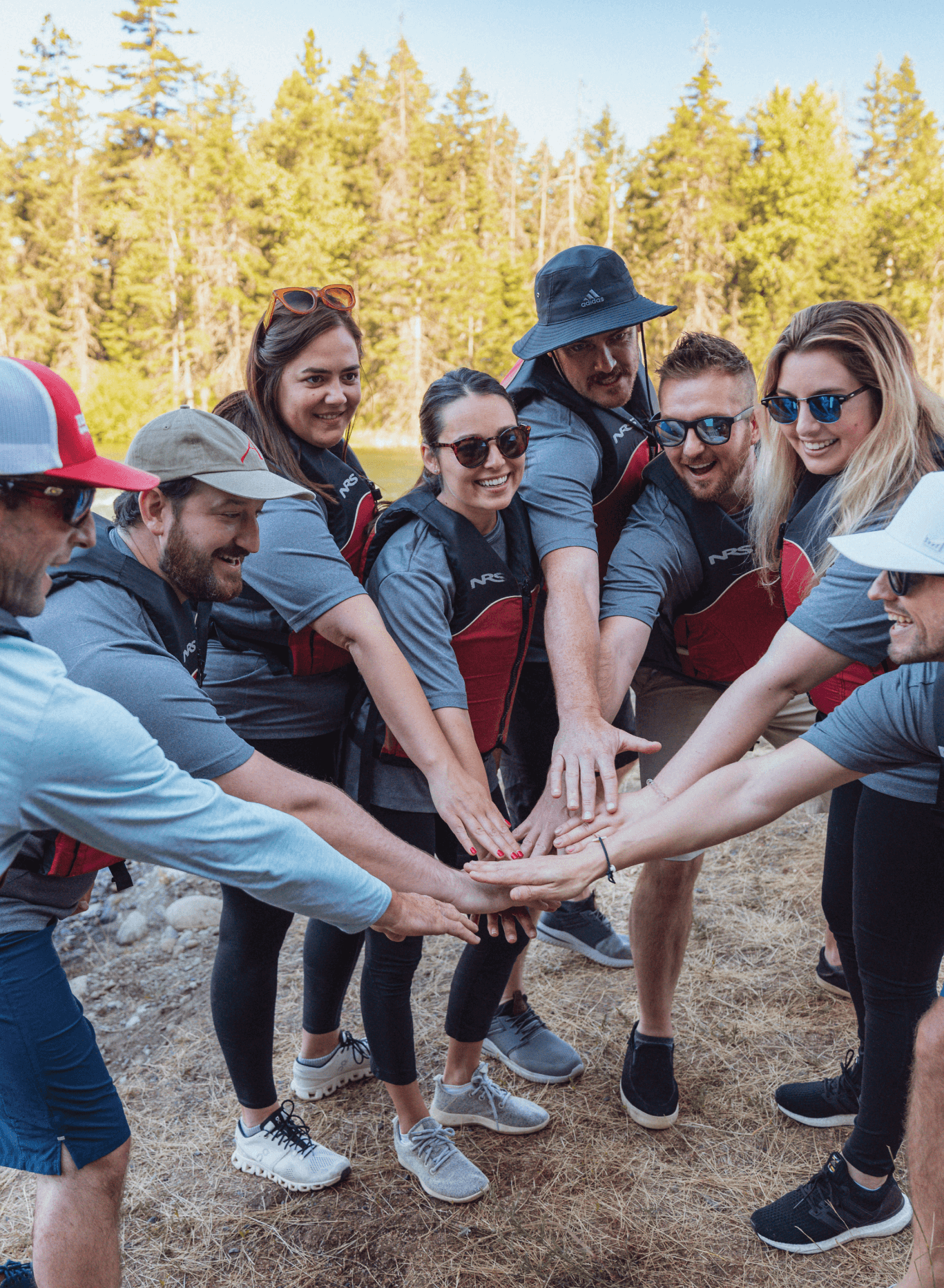 A group of eight people standing outdoors in a semicircle, all extending their hands to the center in a team gesture, with trees and blue sky in the background.