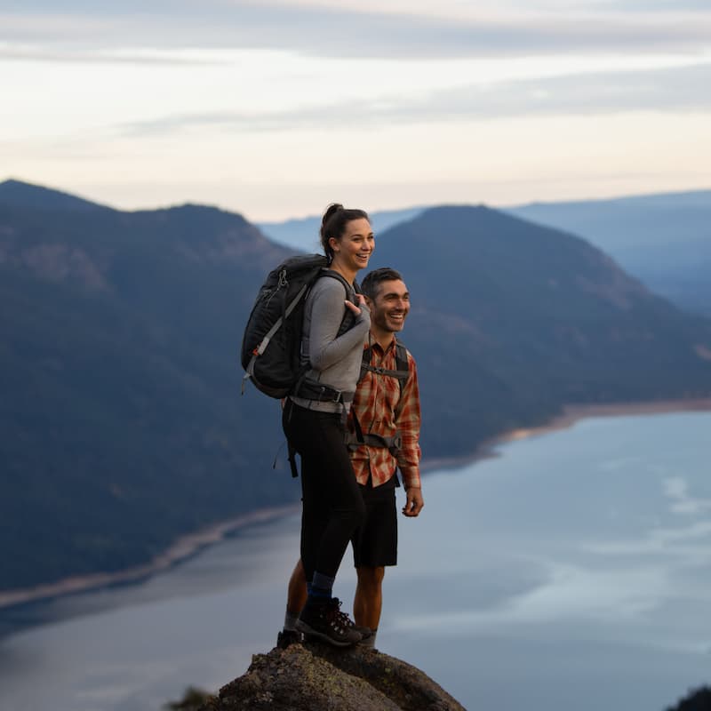 Two people with backpacks stand on a rocky outcrop overlooking a lake and mountain landscape at sunset.