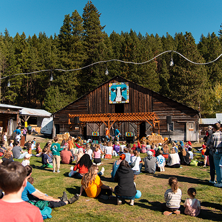 A crowd sits on grass facing a wooden barn stage, watching performers. The scene is outdoors on a sunny day, with tall trees in the background.