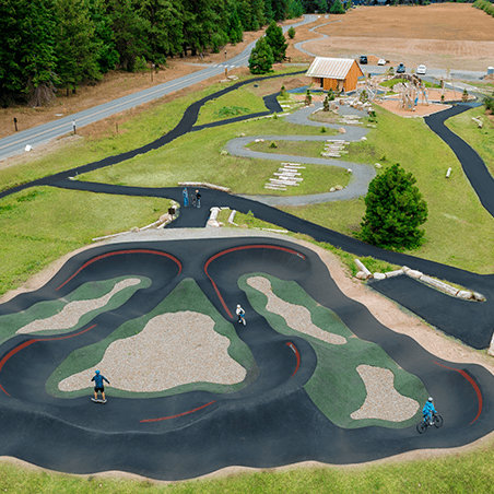 Aerial view of an outdoor paved pump track with people riding bikes and walking, surrounded by grass, trees, and a nearby playground.