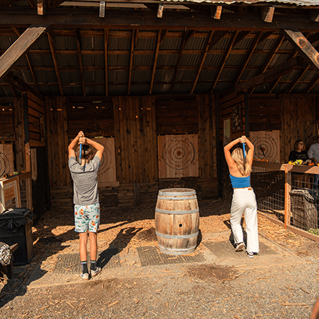 Two people stand side by side, holding axes over their heads, preparing to throw at wooden target boards in an outdoor axe-throwing area.
