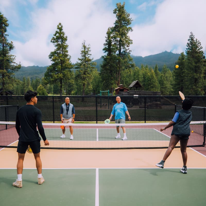 Four people play a game of pickleball on an outdoor court surrounded by trees and mountains under a partly cloudy sky.