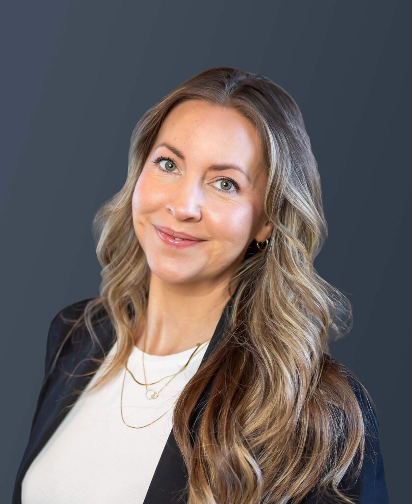 A woman with long, wavy blonde hair, wearing a white top, black blazer, and layered gold necklaces, smiles against a dark gray background.