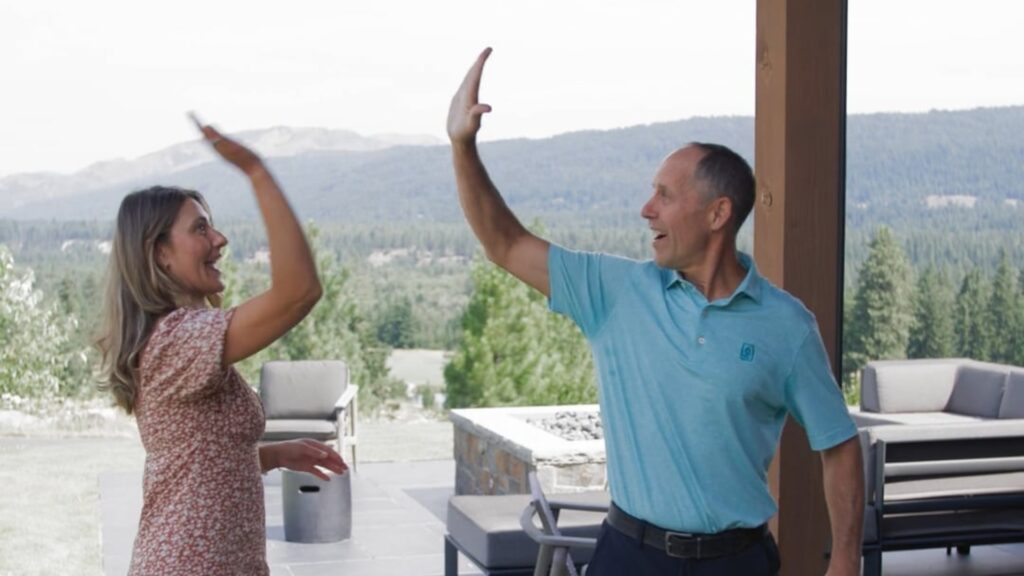 A woman and a man stand outdoors by patio furniture, smiling and giving each other a high five with a forest and mountains in the background.