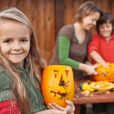 A girl holds a carved pumpkin with plastic spiders, while two people in the background carve another pumpkin on a table outdoors.