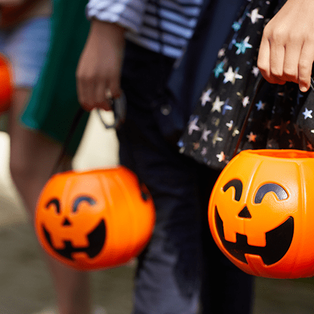 Children holding orange plastic pumpkin buckets while trick-or-treating, dressed in costumes for Halloween.