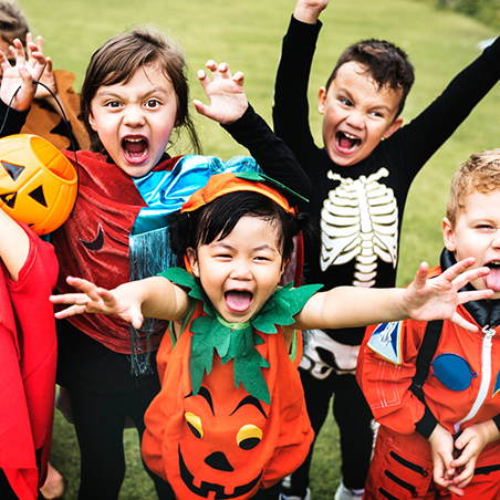 A group of children in Halloween costumes smile and raise their arms enthusiastically while standing outdoors on grass.
