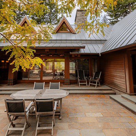 A patio with a round wooden table and six chairs sits outside a wood and metal house, surrounded by trees and autumn leaves on the ground.