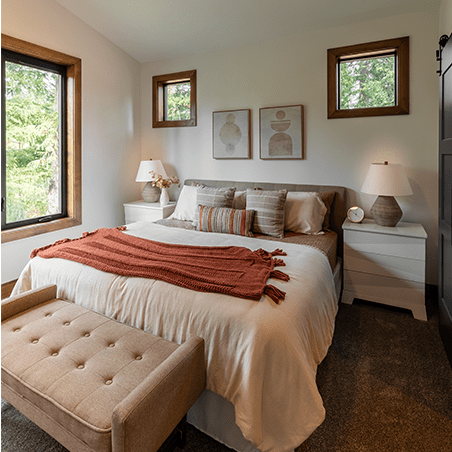 A neatly made bed with white and rust-colored bedding, two side tables with lamps, a bench at the foot, and three framed artworks on the wall in a bright, modern bedroom.