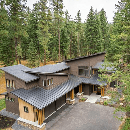 A modern two-story house with a dark sloped roof is surrounded by tall pine trees and features large windows and wood accents.