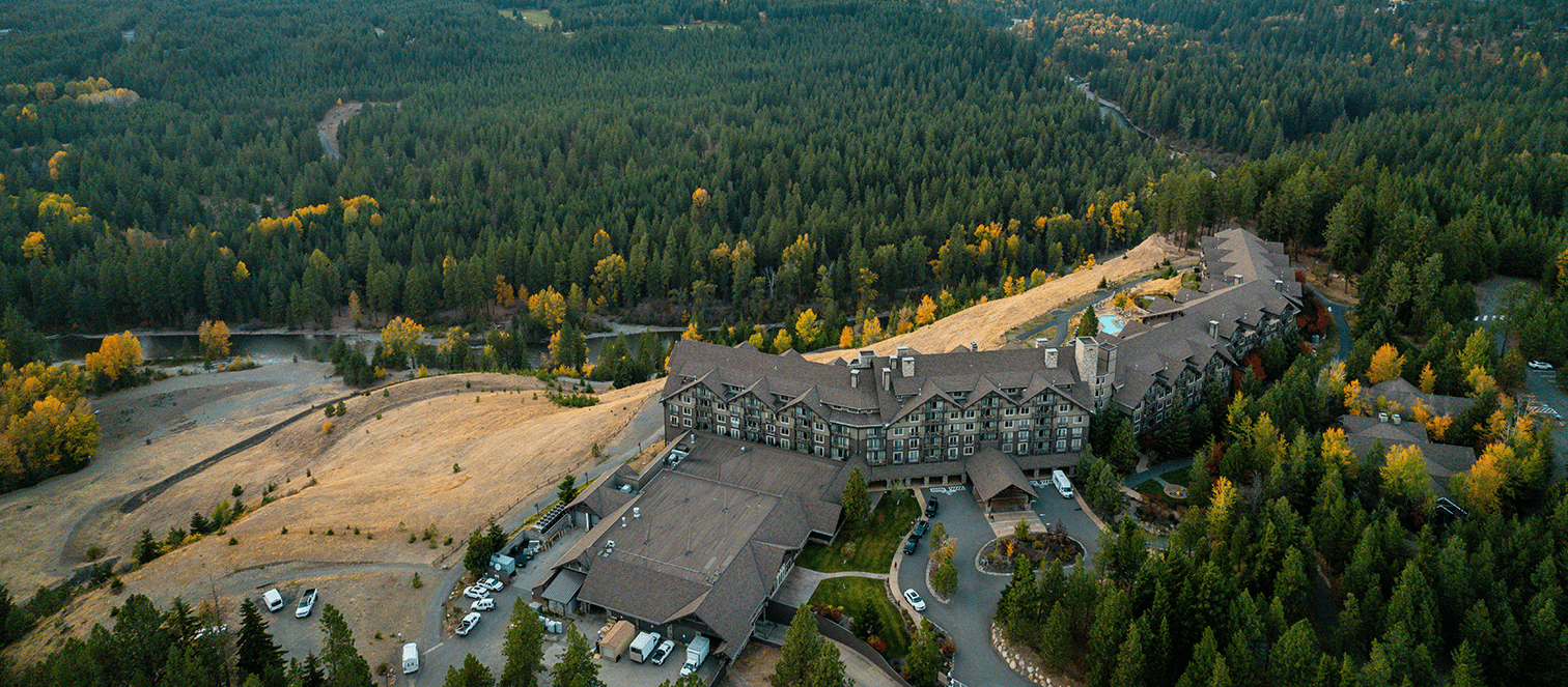 A large lodge-style building surrounded by forested hills and scattered autumn trees, with a winding driveway and several parked cars visible.